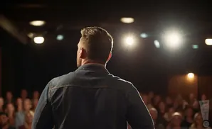 Man in denim jacket on stage facing away from camera, speaking to an audience under spotlights.