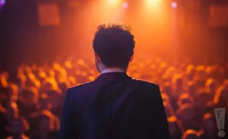 A man in a suit faces a cheering crowd under warm stage lights.