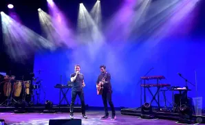 Two musicians perform on a brightly lit stage with blue and purple lighting. One sings into a microphone, the other plays guitar.