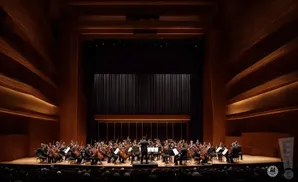 An orchestra performs on stage in a wood-paneled concert hall with warm lighting.