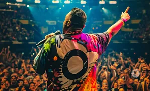 A performer with a colorful shirt sings into a microphone with his arm raised to a cheering crowd.