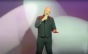 Comedian Bill Burr stands center stage in a black shirt and pants, holding a microphone and gesturing with his right hand.