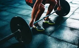 A person in athletic shorts and shoes deadlifts a barbell on a black rubber gym floor.