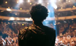 A man with curly hair stands on stage in a decorative jacket, facing a blurred audience under bright lights.