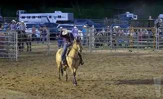 Cowboy on a tan horse in a rodeo arena, with spectators and trailers in the background.