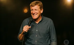 A smiling man in a denim shirt holds a microphone on a dark stage with warm lighting.