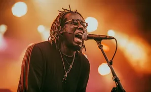 A singer with dreadlocks and glasses sings into a microphone on a stage lit by warm lights.
