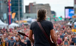 Guitarist with long hair performs for a large crowd at an outdoor festival.
