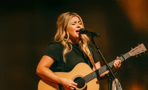 A blonde woman sings into a microphone while playing an acoustic guitar on stage.