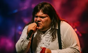 A man with long dark hair sings into a microphone, wearing a white ruffled shirt and black suspenders.