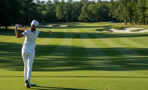 Golfer in white swings club on a manicured golf course with sand traps and trees.