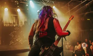 A guitarist performs on stage, her back to the camera, with colorful lighting and a crowd visible.