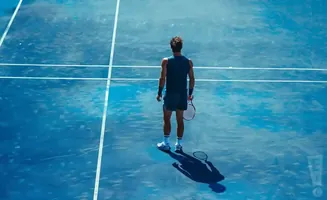 Tennis player on a blue court, holding a racket and casting a shadow.
