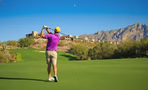 Golfer in purple shirt and yellow hat swings at a golf ball on a sunny day in the desert.