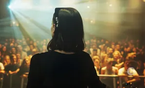 Woman in black shirt and jeweled headband faces away from the camera, looking at a cheering crowd under stage lights.