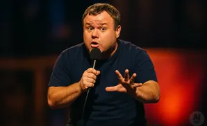 A man with wide eyes and an open mouth speaks into a microphone, gesturing with his hands.