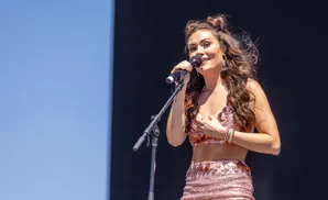 A woman in a sparkly pink outfit sings into a microphone on a stage with a dark background.