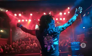 Rock musician on stage with arms raised in front of a cheering crowd, bathed in red and blue stage lights.