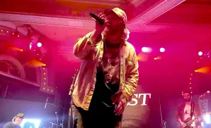 A man in a tan jacket and cap sings into a microphone on a pink-lit stage. Another man plays guitar to his right.