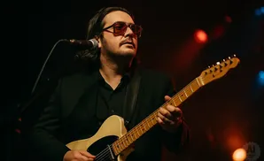 Man in a suit and sunglasses plays a cream-colored electric guitar on stage, with red and blue lights in the background.