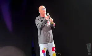 Bald man in a long-sleeved shirt and jeans with colorful patches speaks into a microphone on stage.