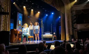A scene from a play with three actresses on stage, a piano, and a bed. An orchestra is in the foreground.