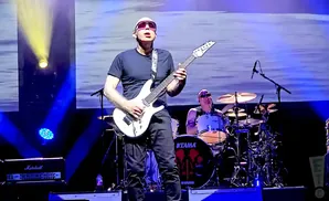 Guitarist Joe Satriani performs on stage with a white guitar, wearing sunglasses, with a drummer behind him.