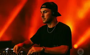 DJ in black t-shirt and cap with headphones, mixing on a console with red-orange lighting.