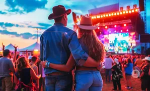 Couple in cowboy hats hug at a brightly lit outdoor concert.