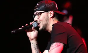 A man with glasses and a backward baseball cap sings into a microphone on stage.