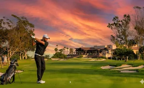 Golfer swings club on a course at sunset, with a clubhouse in the background.