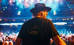 A man in a black hat and t-shirt stands on a stage, facing away from the camera, with a crowd and bright lights behind him.