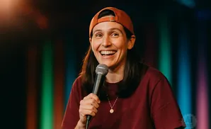 A comedian in a baseball cap and maroon shirt laughs while holding a microphone.