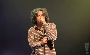 A person with curly hair sings into a microphone while wearing a brown and white striped shirt.