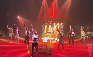 Circus performers in costume point up during a show on a red stage under red lights.