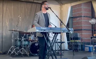 A man in a grey suit jacket sings into a microphone while playing a Roland Jupiter-50 keyboard at an outdoor concert.