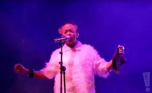 A person in a fluffy white coat and bear ears sings into a microphone on a purple-lit stage.