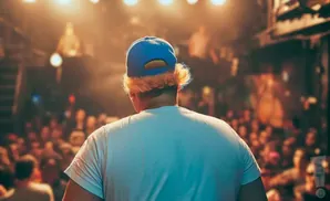 Person with blonde hair peeking from under a blue baseball cap on stage facing a blurry crowd.