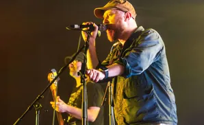 A man with a beard and a brown cap sings into a microphone on stage, with another musician in the background.