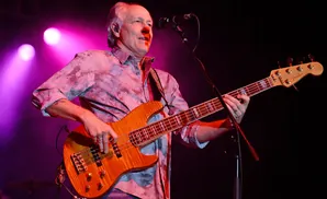 A man plays a Fender bass guitar on stage with purple lighting.