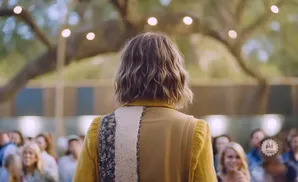 Person with wavy blonde hair facing an audience, wearing a yellow sweater and a tan vest with floral and furry accents.