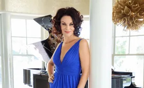 Woman in a blue dress leans against a white pillar near a grand piano.