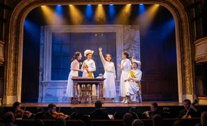 Five women in white dresses perform on stage with an orchestra below.