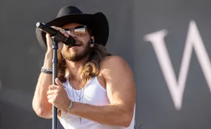 Man in a cowboy hat and white tank top sings into a microphone, wearing sunglasses that reflect a crowd.
