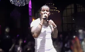 A male rapper in a white t-shirt and chains performs on stage with a crowd in the background.