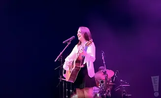 A woman sings and plays an acoustic guitar on stage, bathed in purple light.