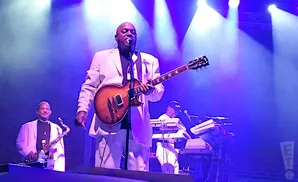 A band performs on stage with a guitarist in a white suit singing, a saxophonist to his left, and a keyboardist behind him.