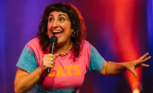A comedian with dark, curly hair laughs into a microphone while wearing a tie-dye shirt with 