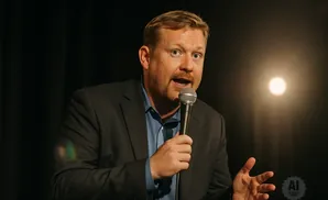 A man in a suit speaks into a microphone with a spotlight behind him.