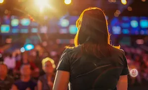 Woman with auburn hair in a dark shirt facing a bright stage with a blurred audience.